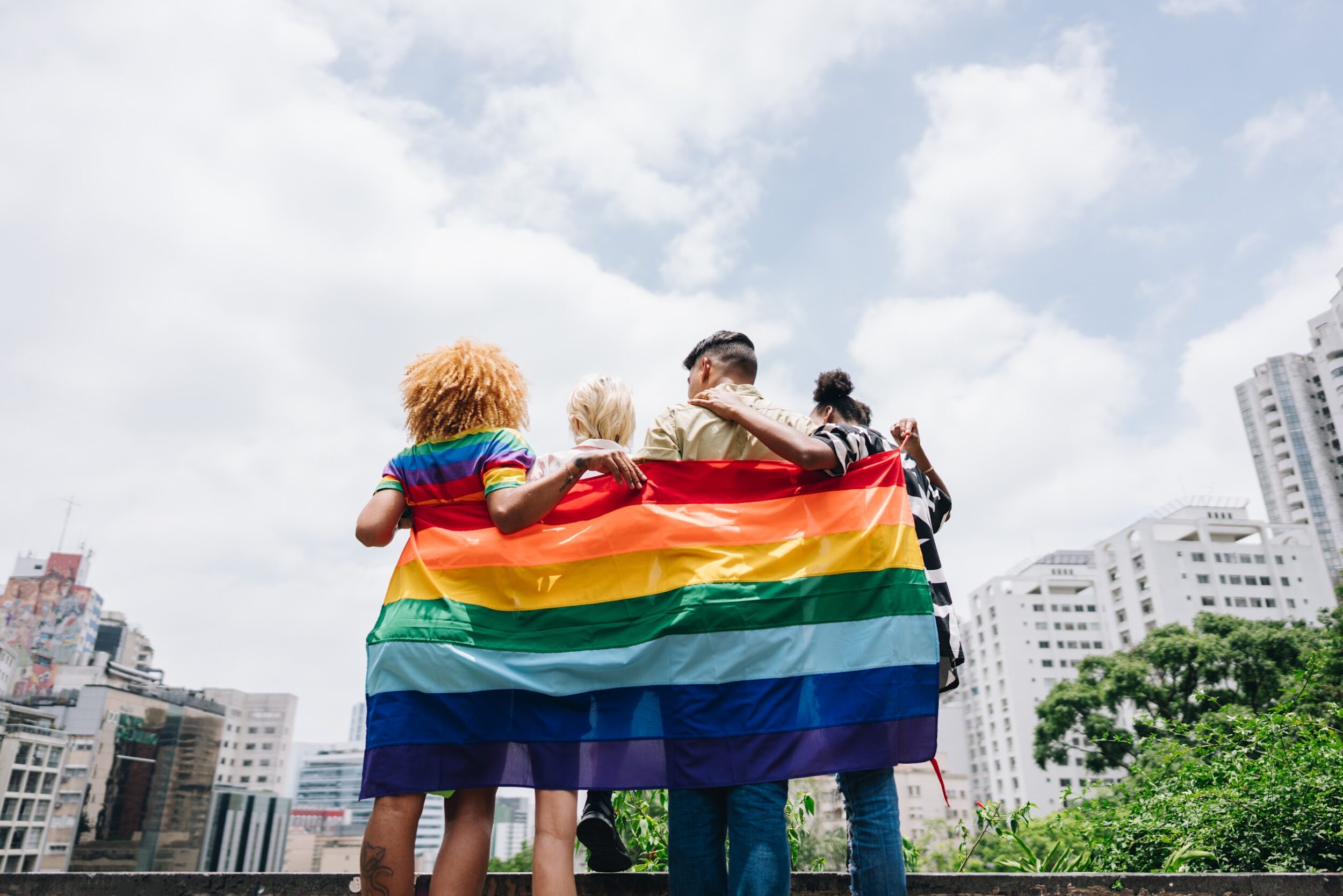 Lgbtqia+ friends embracing and holding a rainbow flag outdoors unnamed 6 scaled